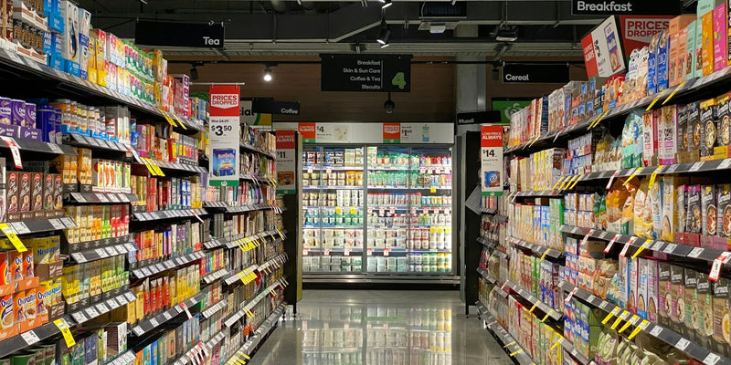 Supermarket aisle with stocked shelving leading to glass-door display fridges at the back of the store